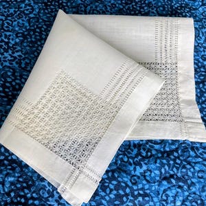 May include: Two folded white linen napkins with intricate openwork and embroidered borders. The napkins are displayed on a blue patterned fabric, showcasing the delicate details of the textile.
