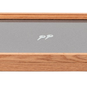 May include: A rectangular wooden display case with a glass top and a gray interior. Two small, white keys are inside. The case has brass-colored latches and a light brown wood finish.