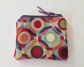 May include: A small, square coin purse with a colorful geometric pattern in red, orange, green, blue, and cream. The purse has a purple zipper and a matching pull. The purse is made of woven fabric.