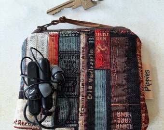 May include: A small, rectangular zippered pouch with a woven book design in red, black, and blue. The pouch has a brown leather pull and a small tag that says "Pippies". Black earbuds and a key are next to the pouch.