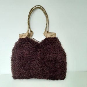 Peut inclure: Un sac fourre-tout marron foncé en peluche avec un extérieur texturé. Le sac est doté de poignées beiges avec un motif cousu noir et or. Les poignées sont conçues pour porter le sac.
