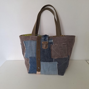 Op de afbeelding: Een tote bag gemaakt van patchwork denim in diverse tinten blauw en bruin. De tas is voorzien van twee olijfgroene schouderbanden, een centrale verticale olijfgroene strook met metalen ringen en een sluiting met een bronskleurige metalen gesp.