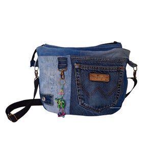 Könnte beinhalten: Jeans-Umhängetasche mit Patchwork-Design. Die Tasche hat ein Wrangler-Etikett und einen dekorativen Anhänger. Sie hat einen schwarzen, verstellbaren Riemen und Metallbeschläge. Der Jeansstoff ist eine Mischung aus hellen und dunklen Blautönen.