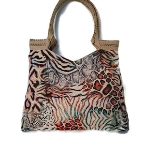 Op de afbeelding: Een tote bag met een uniek dierenprint ontwerp. De tas heeft een mix van zebra strepen, reptielschubben en luipaardvlekken in bruin, beige, rood en groen. De handvatten zijn bruin.