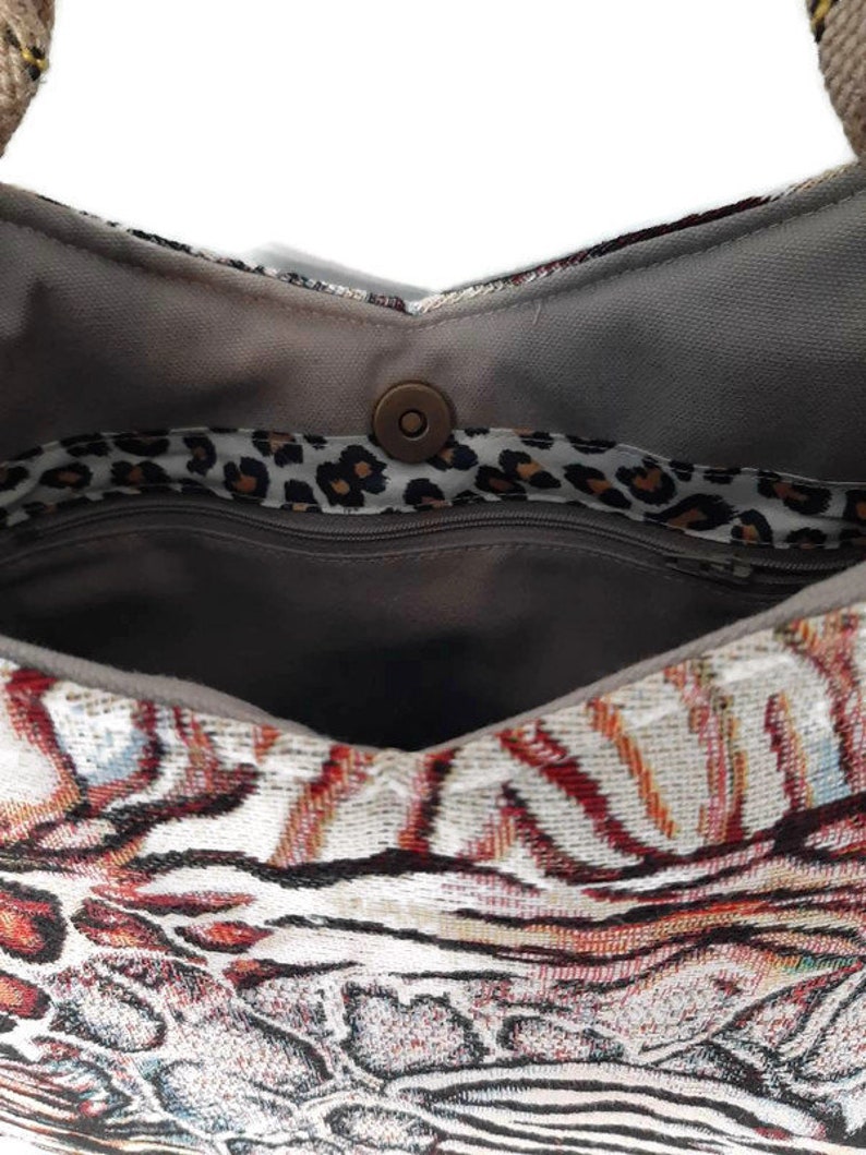 Op de afbeelding: Close-up van een patroon draagtas. De tas heeft een geweven ontwerp met rode, bruine en witte tinten. De binnenkant is grijs met een luipaardprint voering en een magnetische sluiting.