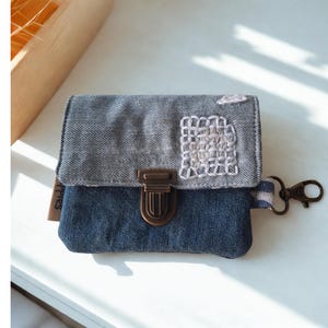 Op de afbeelding: Een tweekleurige denim portemonnee met klep en metalen sluiting. Het bovenste gedeelte is grijs, het onderste blauw. Het heeft een wit geborduurd detail en een gestreepte band met een karabijnhaak. Ongeveer 15 cm breed.