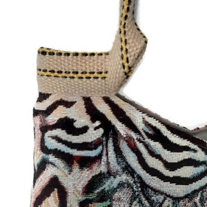 Op de afbeelding: Detail van een geweven draagtas met een patroon. De tas heeft een beige band met gele en zwarte stiksels. De hoofdbody van de tas toont een kleurrijk patroon met zwarte, witte en blauwe tinten.