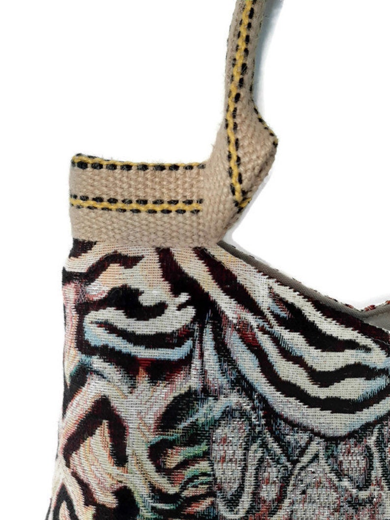 Op de afbeelding: Detail van een geweven draagtas met een patroon. De tas heeft een beige band met gele en zwarte stiksels. De hoofdbody van de tas toont een kleurrijk patroon met zwarte, witte en blauwe tinten.