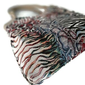Op de afbeelding: Een handtas met patroon en een geweven ontwerp. De tas heeft een mix van dierenprints, waaronder zebra strepen en luipaardvlekken, in tinten zwart, bruin, rood en wit. De handgreep is lichtbruin.