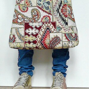 Op de afbeelding: Een grote tote bag met een patchwork ontwerp met paisley, geometrische en tribale patronen in rood, cr&egrave;me en zwart. De tas wordt voor een persoon gehouden die een blauwe spijkerbroek en sneakers draagt. De tekst "same size, other bag" staat onderaan.