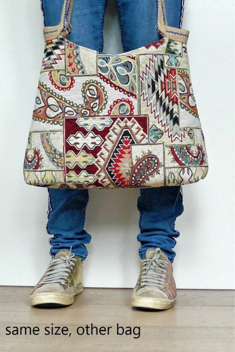 Op de afbeelding: Een grote tote bag met een patchwork ontwerp met paisley, geometrische en tribale patronen in rood, cr&egrave;me en zwart. De tas wordt voor een persoon gehouden die een blauwe spijkerbroek en sneakers draagt. De tekst "same size, other bag" staat onderaan.