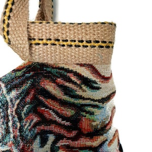 Op de afbeelding: Close-up van een geweven draagtas met een kleurrijk, abstract patroon. De tas heeft een bruine jute handgreep en afwerking met gele en zwarte stiksels. De hoofdbody van de tas toont een mix van rode, blauwe en zwarte tinten.