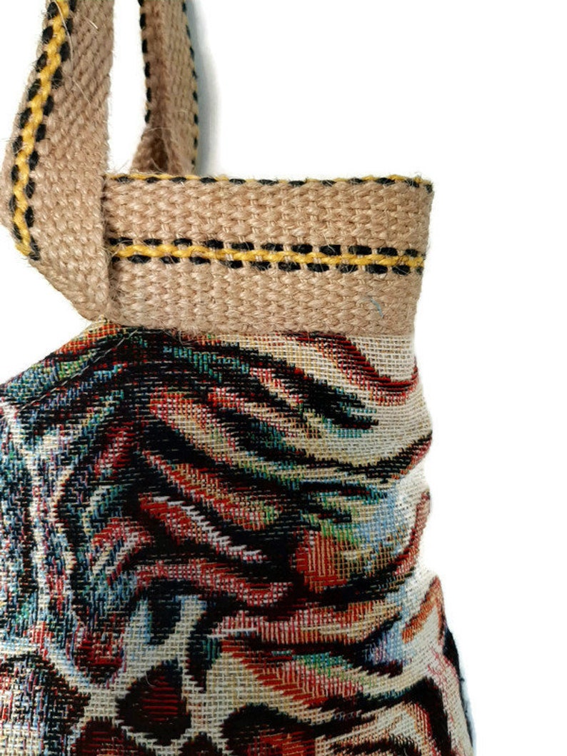 Op de afbeelding: Close-up van een geweven draagtas met een kleurrijk, abstract patroon. De tas heeft een bruine jute handgreep en afwerking met gele en zwarte stiksels. De hoofdbody van de tas toont een mix van rode, blauwe en zwarte tinten.