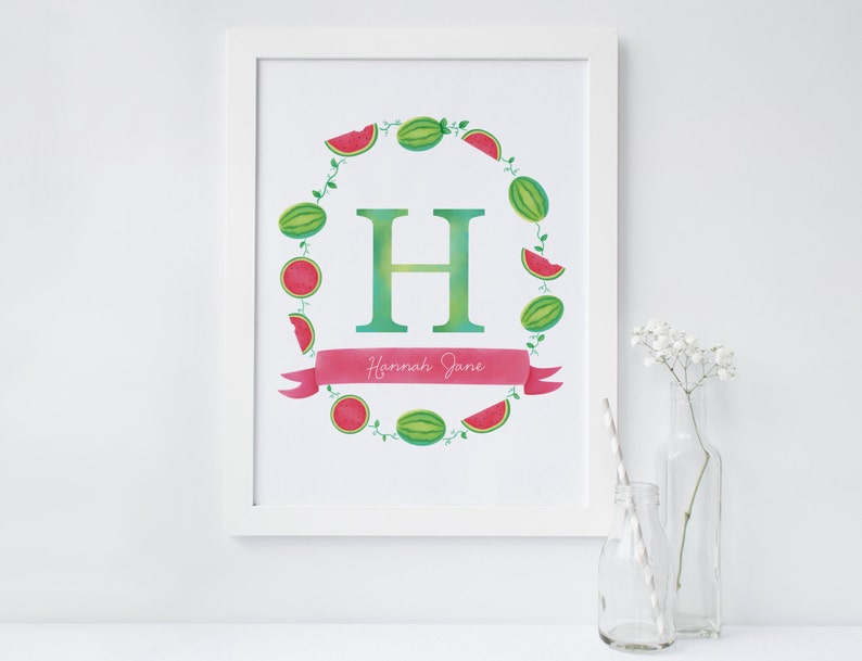 May include: A white framed print with a watercolor illustration of a green letter H surrounded by a wreath of watermelon slices. The wreath is on a white background with a pink ribbon that says "Hannah Jane".