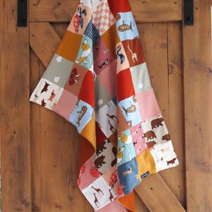 Peut inclure: Une courtepointe patchwork aux motifs animaliers et géométriques variés, dans des tons de rouge, orange, bleu, marron et rose. La courtepointe est suspendue à une porte en bois. Elle présente des ours, des chiens, des girafes et d'autres motifs.