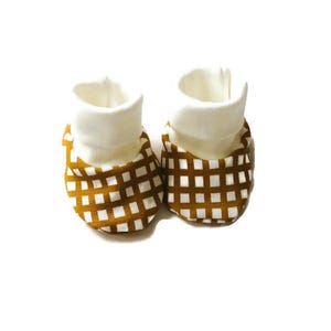 Puede incluir: Un par de patucos de bebé con interior y puño blancos. El exterior presenta un estampado de cuadros marrones y blancos. Los patucos están diseñados para mantener los pies del bebé calientes y cómodos.