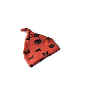 Puede incluir: Un gorro de bebé de color óxido con un nudo en la parte superior. El gorro está decorado con un patrón de origami negro de animales y mariposas.