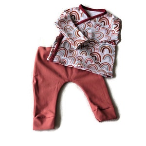 Peut inclure: Un ensemble de vêtements pour bébé composé d'un haut à manches longues et d'un pantalon. Le haut est blanc avec un motif arc-en-ciel dans les tons de brun, rose et orange. Le pantalon est de couleur rouille unie.