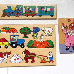 Peut inclure: Trois puzzles en bois à boutons avec des illustrations colorées : un train avec des passagers, une scène de ferme avec un tracteur, des animaux et une personne, et un clown jonglant. Chaque pièce de puzzle comporte un petit bouton en bois.