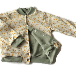 Puede incluir: Una chaqueta bomber para bebé de color verde y amarillo con estampado floral y cierre de presión.