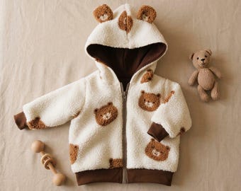 Può includere: Una giacca a maniche lunghe con cerniera in materiale peluche color crema, dotata di cappuccio con due piccole orecchie da orso. La giacca è decorata con toppe a forma di muso d'orso marrone e presenta polsini e orlo marrone scuro.