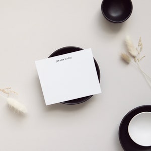May include: A white card with the text "Just a note to say" on a black saucer. The card is on a light gray background with two small dried flower stems.