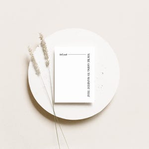 May include: A minimalist composition featuring a white card with the text "You're going to manifest that" and "Dell yeah" on a white circular plate. Dried wheat stalks add a natural touch to the scene, set against a neutral background.