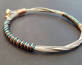May include: A handmade bracelet made from silver-coloured guitar strings. The bracelet has copper and teal-coloured wire accents, with a gold-coloured clasp. This unique jewellery piece is a stylish accessory.