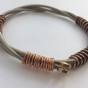 May include: A bracelet made from repurposed guitar strings. The bracelet is made of silver, copper, and black strings, and has a gold clasp.