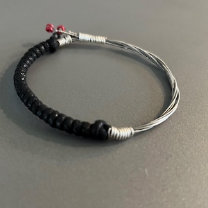 Puede incluir: Una pulsera hecha a mano con cordón negro y alambre de metal plateado. La pulsera presenta una sección negra tejida y un bucle de alambre plateado. Pequeñas cuentas rojas acentúan el diseño.