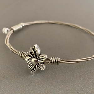 Könnte beinhalten: Ein silbernes Armband aus Gitarrensaiten mit einem silbernen Blumenanhänger.