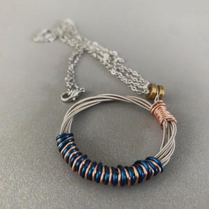 Può includere: Una collana d'argento con un ciondolo circolare fatto di corde di chitarra. Il ciondolo presenta accenti blu e color rame. La collana è esposta su una superficie grigia.