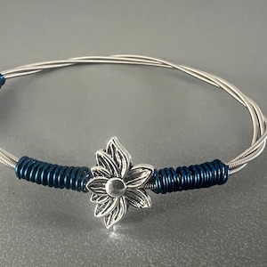 Könnte beinhalten: Ein silberfarbenes Armband aus gewickeltem Draht mit einem silbernen Blumenanhänger in der Mitte. Der Draht ist in Abständen mit dunkelblauem Material umwickelt. Ein einzigartiges Schmuckstück.