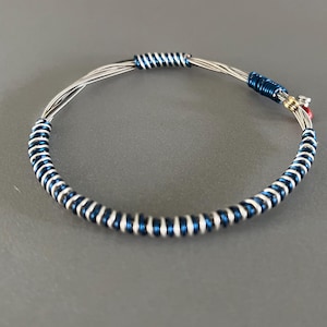 Può includere: Un braccialetto fatto a mano con fili metallici color argento, avvolti con bande alternate di argento e blu scuro. Il braccialetto ha un design circolare con una chiusura e piccoli accenti rossi e dorati.