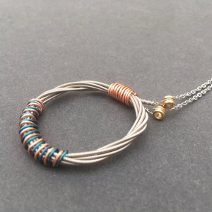May include: A handmade necklace featuring a circular pendant crafted from repurposed guitar strings. The pendant is accented with copper and teal wire wraps, suspended from a silver chain. The design showcases a unique, artistic aesthetic.