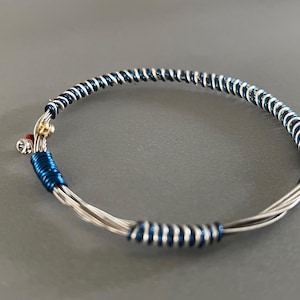 Peut inclure: Bracelet argenté en fil métallique enroulé, avec des détails bleu foncé et argenté. Le bracelet présente un motif en spirale, une petite perle dorée et un accent rouge. Design simple et élégant.