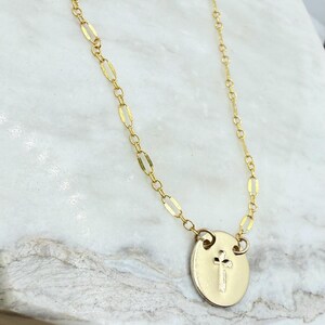 May include: A gold-colored necklace with a delicate chain and a circular pendant. The pendant features a small cross design. The necklace is displayed on a white and gray marble surface.