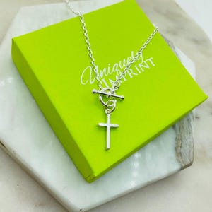 May include: A silver-colored necklace with a cross pendant, displayed on a bright green box. The box has the words "Unique Print" printed on it in a cursive font. The necklace is on a white surface.