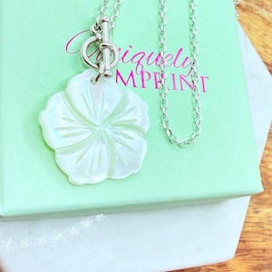 May include: A silver necklace with a white, flower-shaped pendant. The pendant has detailed petal carvings. The necklace is displayed on a light green box with the text "Iquely Mprint". The background is a wooden surface.