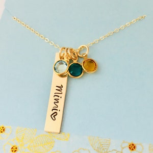 May include: Gold necklace with a bar charm engraved with the word "Minnie" and a heart. The necklace has three small round charms with blue, green, and yellow gemstones.