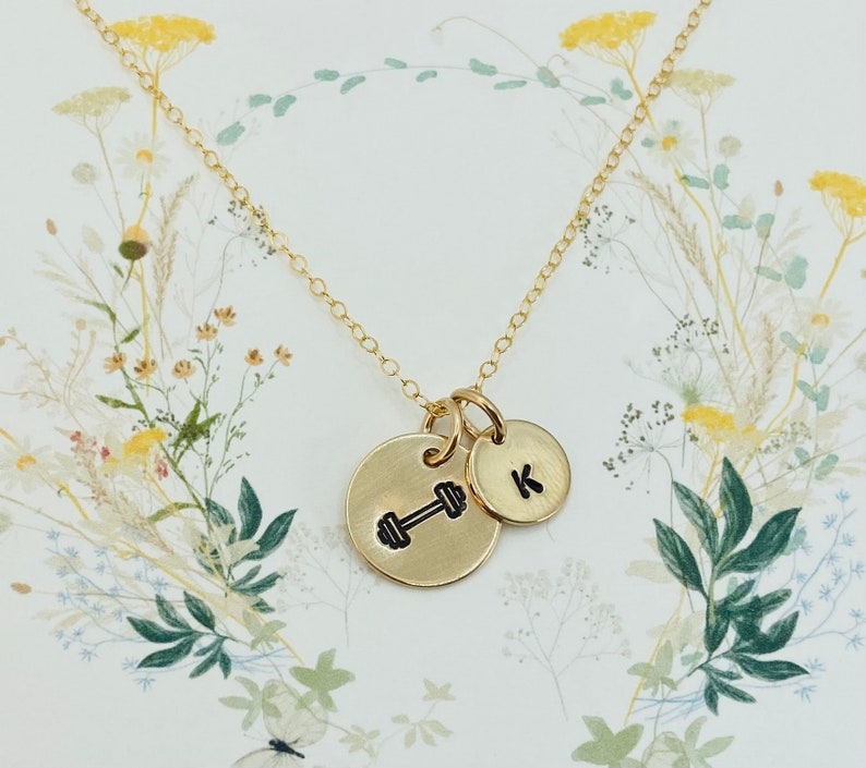 May include: Gold necklace with two charms. One charm is a dumbbell and the other charm is a letter K.