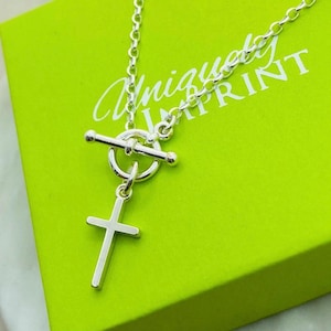 May include: A silver-toned necklace featuring a toggle clasp and a simple cross pendant. The chain is composed of round links, and the toggle mechanism includes a circular ring and a bar. The jewelry is displayed against a bright lime green box.