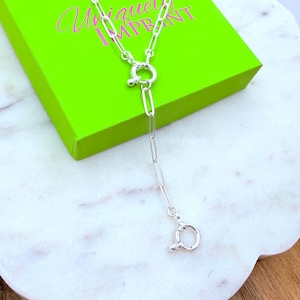 May include: A silver-colored necklace with a unique design. The necklace features a long, rectangular link chain with a circular clasp. The necklace is displayed on a white marble surface, with a bright green box in the background that has the text "Uniquely Imprint".