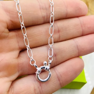 May include: A silver necklace with an oval link chain and a round clasp. The chain links have a textured pattern. The clasp is a circular design with a small ball on top. The necklace is displayed against a neutral background.