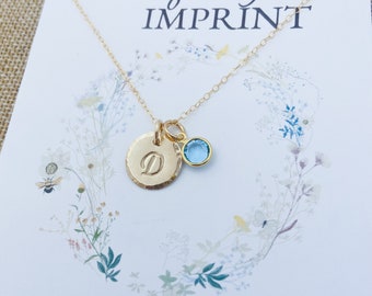 May include: A gold chain necklace featuring a round, hammered gold charm stamped with the letter D and a small, round, light blue crystal charm. The necklace is displayed on a white card with the text Uniquely IMPRINT and floral art.