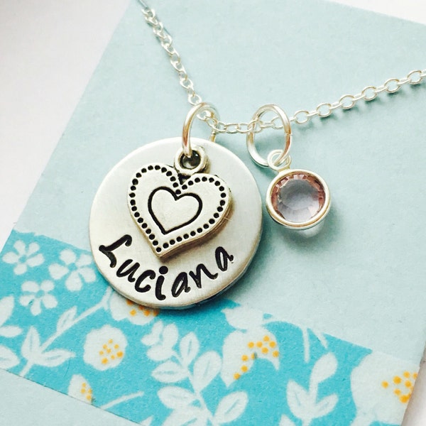 May include: A silver necklace with a pendant featuring a heart-shaped charm and the name "Luciana" engraved on a round disc. A small, round, pink gemstone charm hangs next to the pendant. The necklace is displayed on a light blue card.