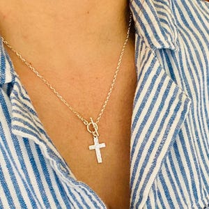 May include: Silver cross pendant necklace with a textured finish, suspended from a delicate chain. The necklace features a toggle clasp and is displayed against a blue and white striped shirt.
