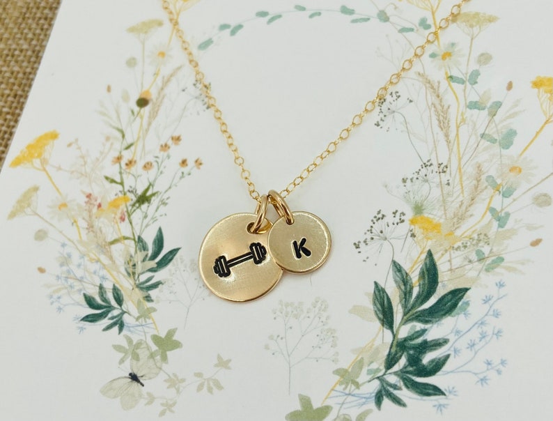 May include: A gold necklace with two charms. One charm is a small circle with the letter "K" on it. The other charm is a larger circle with a dumbbell symbol on it.
