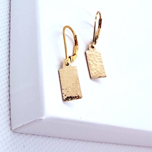May include: A pair of gold-toned drop earrings featuring rectangular pendants with a hammered, textured surface. Each pendant is suspended from a hinged lever-back closure, creating a dangling effect with a metallic finish and a distinct, uneven surface pattern.