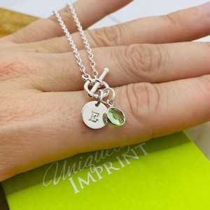 Peut inclure: Un collier en argent avec un fermoir à bascule, orné de deux breloques : un disque rond gravé de la lettre "E" et une petite pierre précieuse ovale vert clair. Le collier est présenté sur une main, avec une boîte verte en arrière-plan.